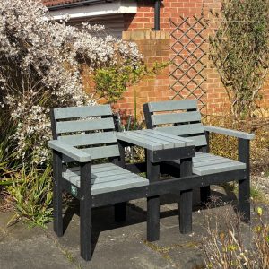 Outdoor seating area with gray wooden benches placed on a black platform, set against a brick wall with a lattice trellis and white flowering shrub to the left.