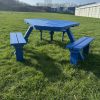 Blue-painted wooden picnic table with attached benches on a grassy field, industrial buildings visible on the horizon under a clear sky.