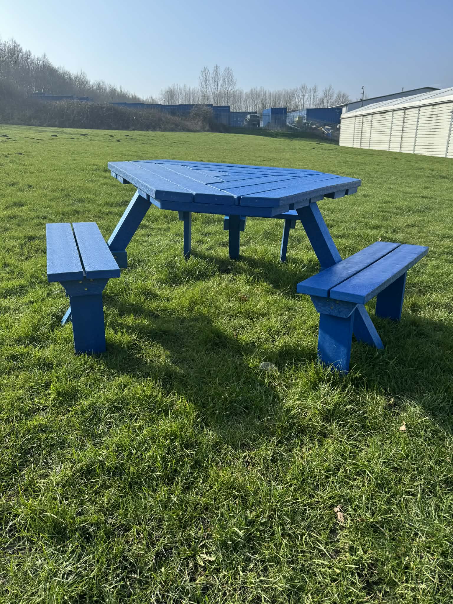 Blue-painted wooden picnic table with attached benches on a grassy field, industrial buildings visible on the horizon under a clear sky.