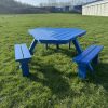 Blue wooden picnic table with benches sits on a grassy field under a clear sky, industrial buildings visible in the background.