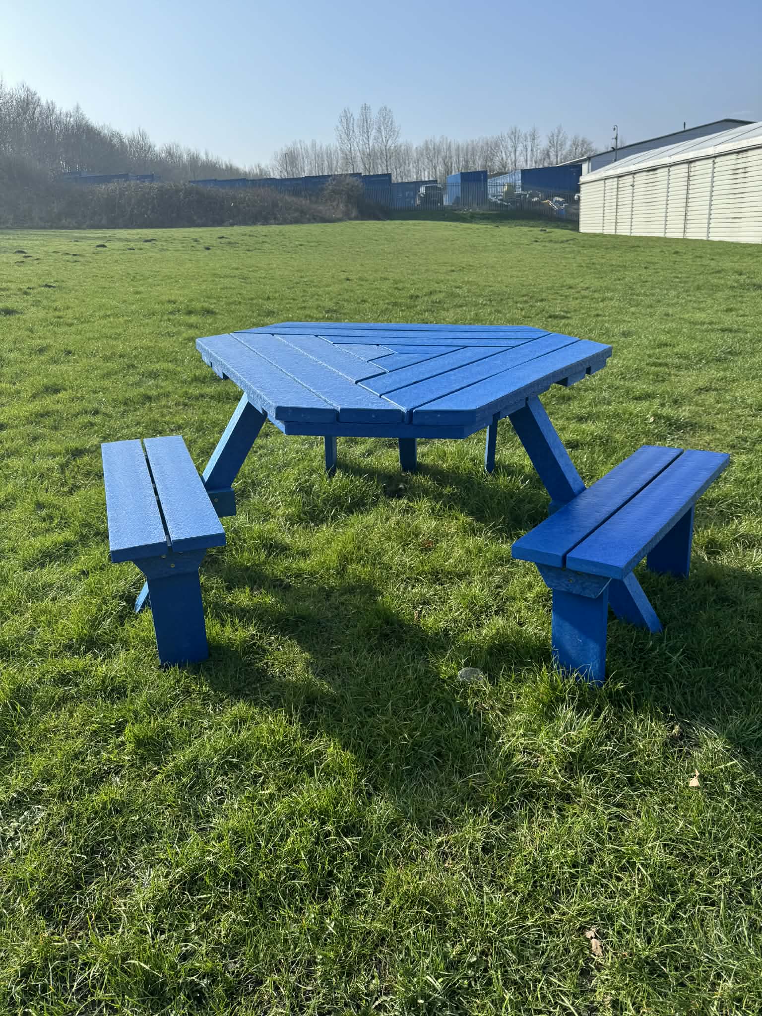 Blue wooden picnic table with benches sits on a grassy field under a clear sky, industrial buildings visible in the background.