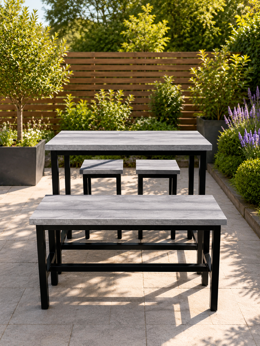 Outdoor gray wooden dining table with matching benches in a sunlit garden patio, lavender and greenery in the background.